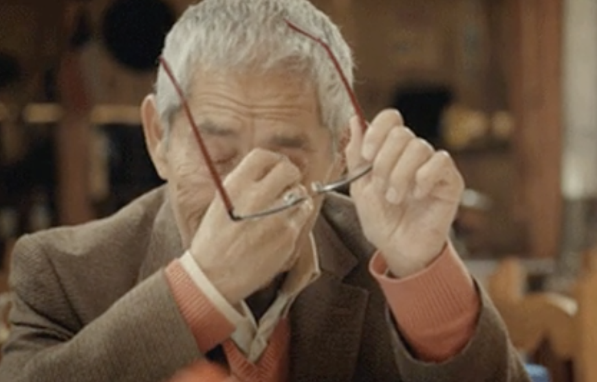 An older man in a sweater pulls off his glasses and wipes his wet eyes