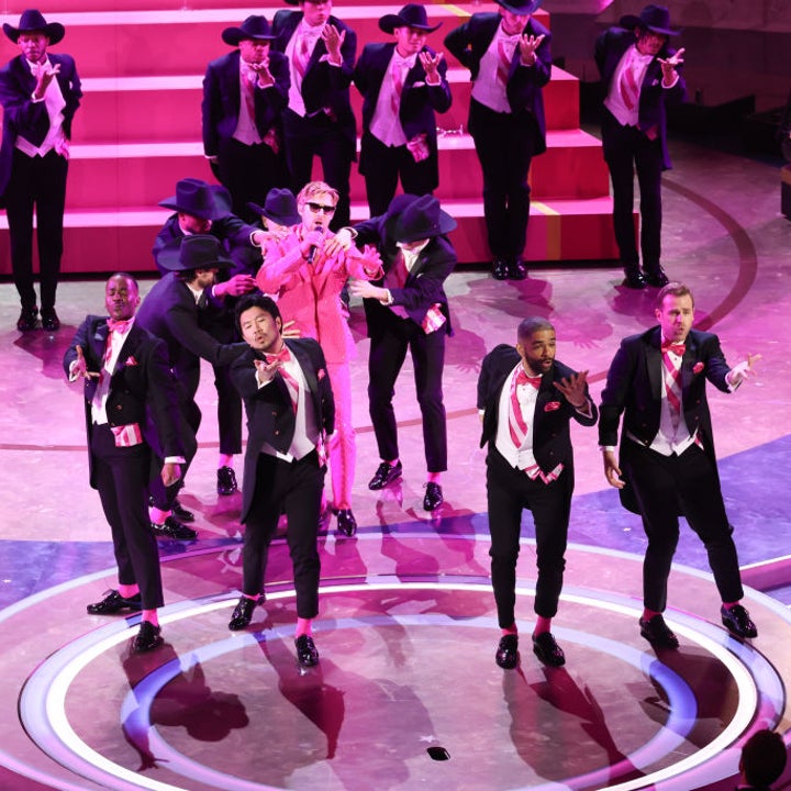 Performers on stage in a dance routine with a central figure in pink, surrounded by others in formal black attire
