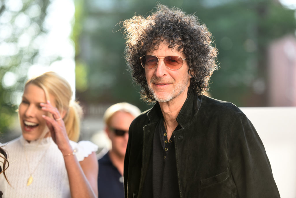Howard Stern wearing a velvet jacket and sunglasses, with a woman laughing beside him