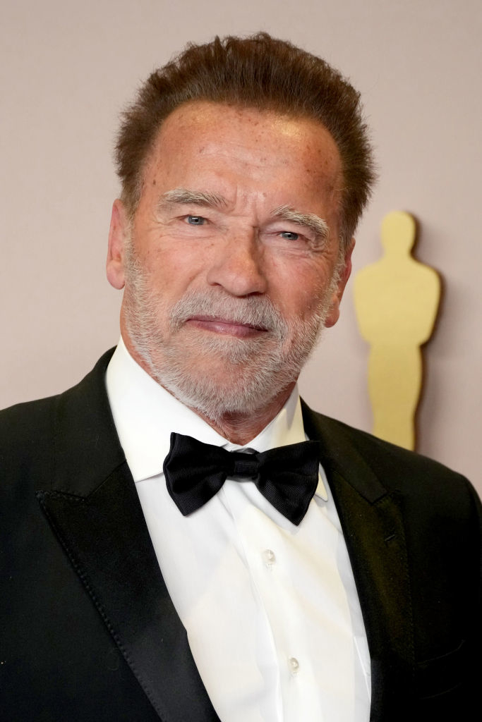Arnold Schwarzenegger wearing a black tuxedo with a bow tie, standing in front of an Oscar statue
