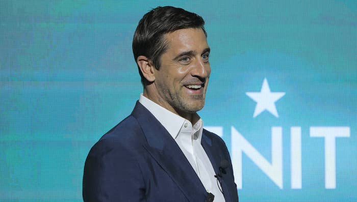 Aaron Rodgers in a blazer and shirt smiling onstage at a sports event