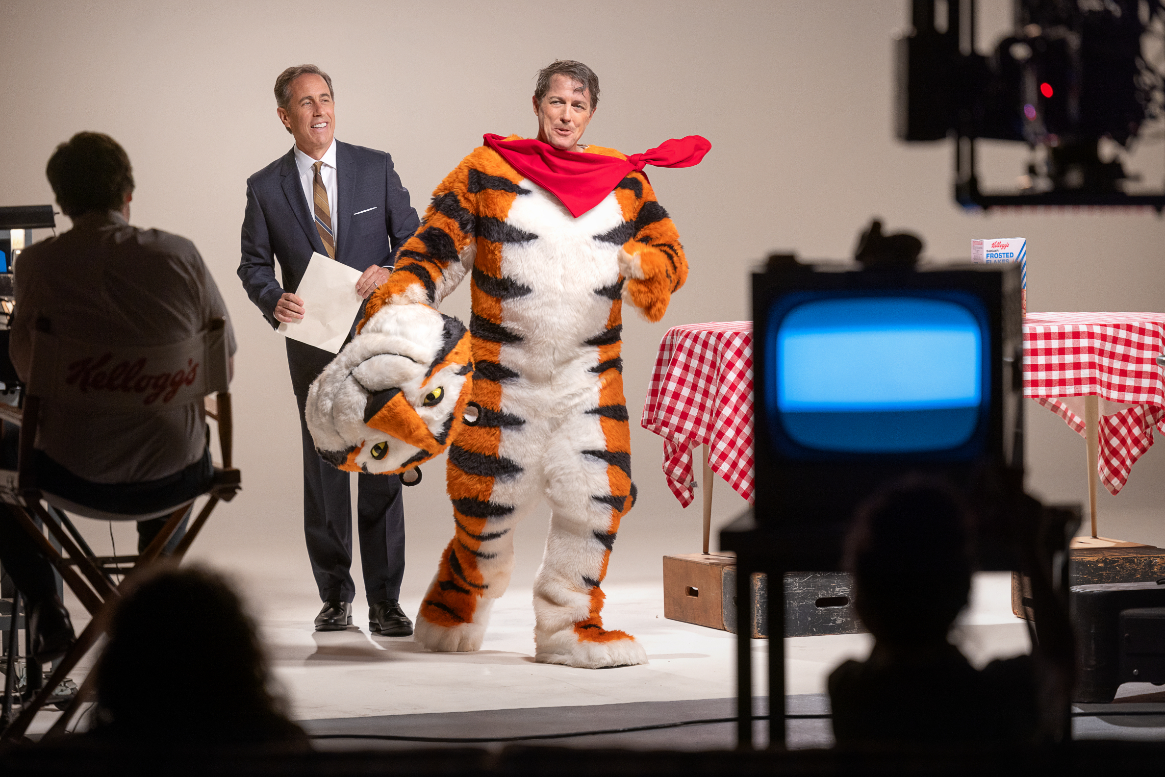Bill Gates in a tiger costume with a red scarf on a set, being interviewed by a man in a suit