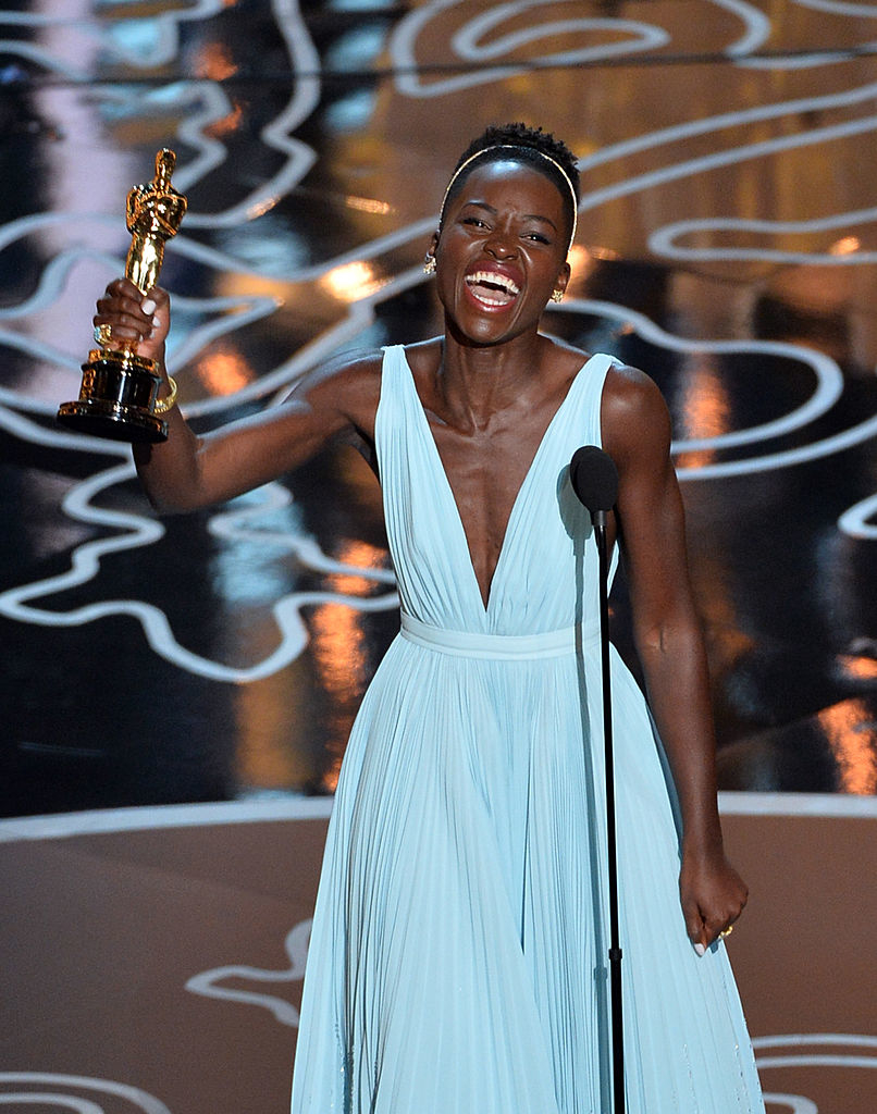 Lupita Nyong&#x27;o in a v-neck gown, joyfully holding an Oscar at the award ceremony