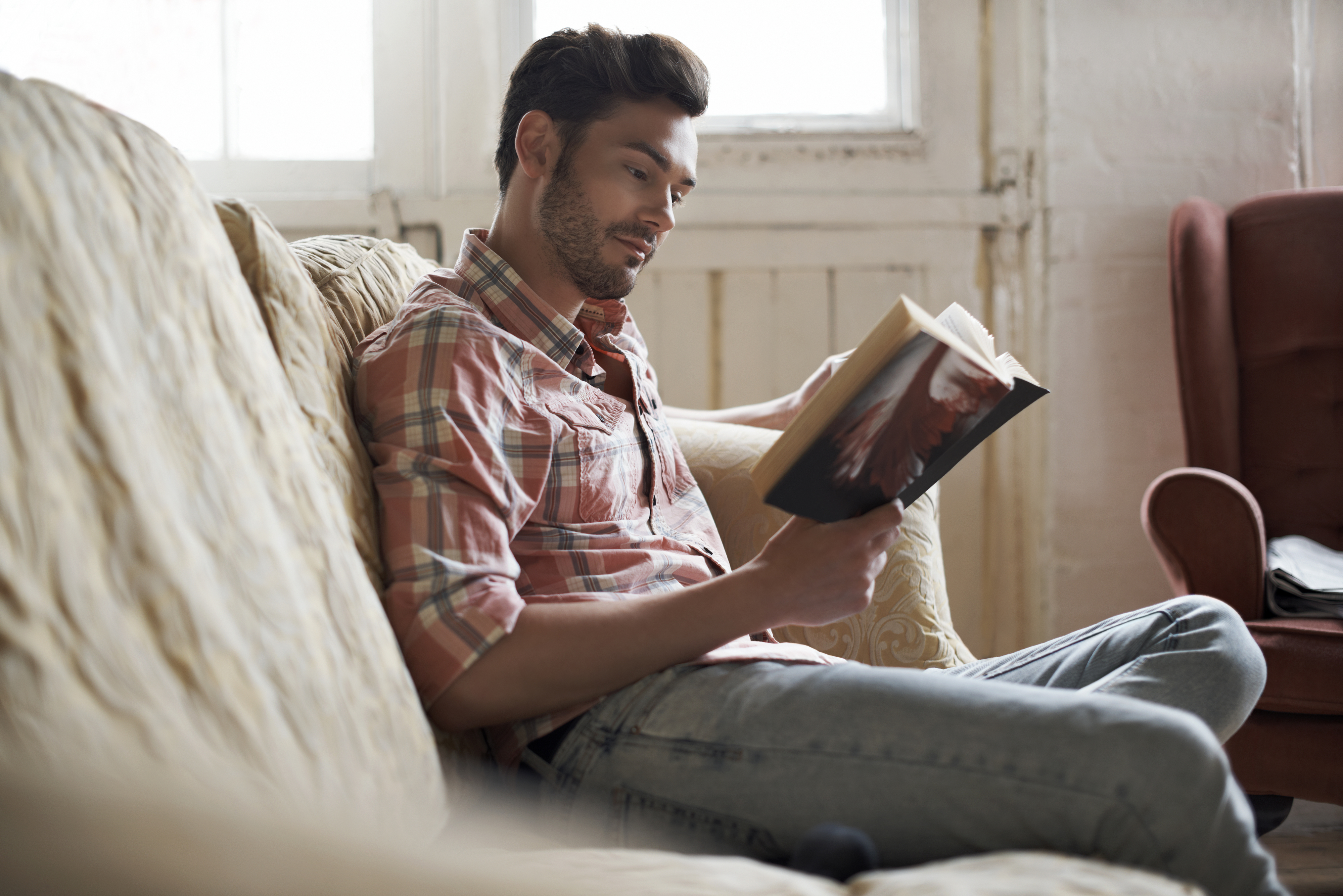 Man in casual shirt sitting on a couch, reading a book with interest
