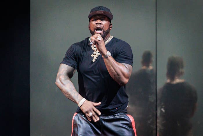 A performer on stage singing into a microphone, wearing a black tee, hat, and chain