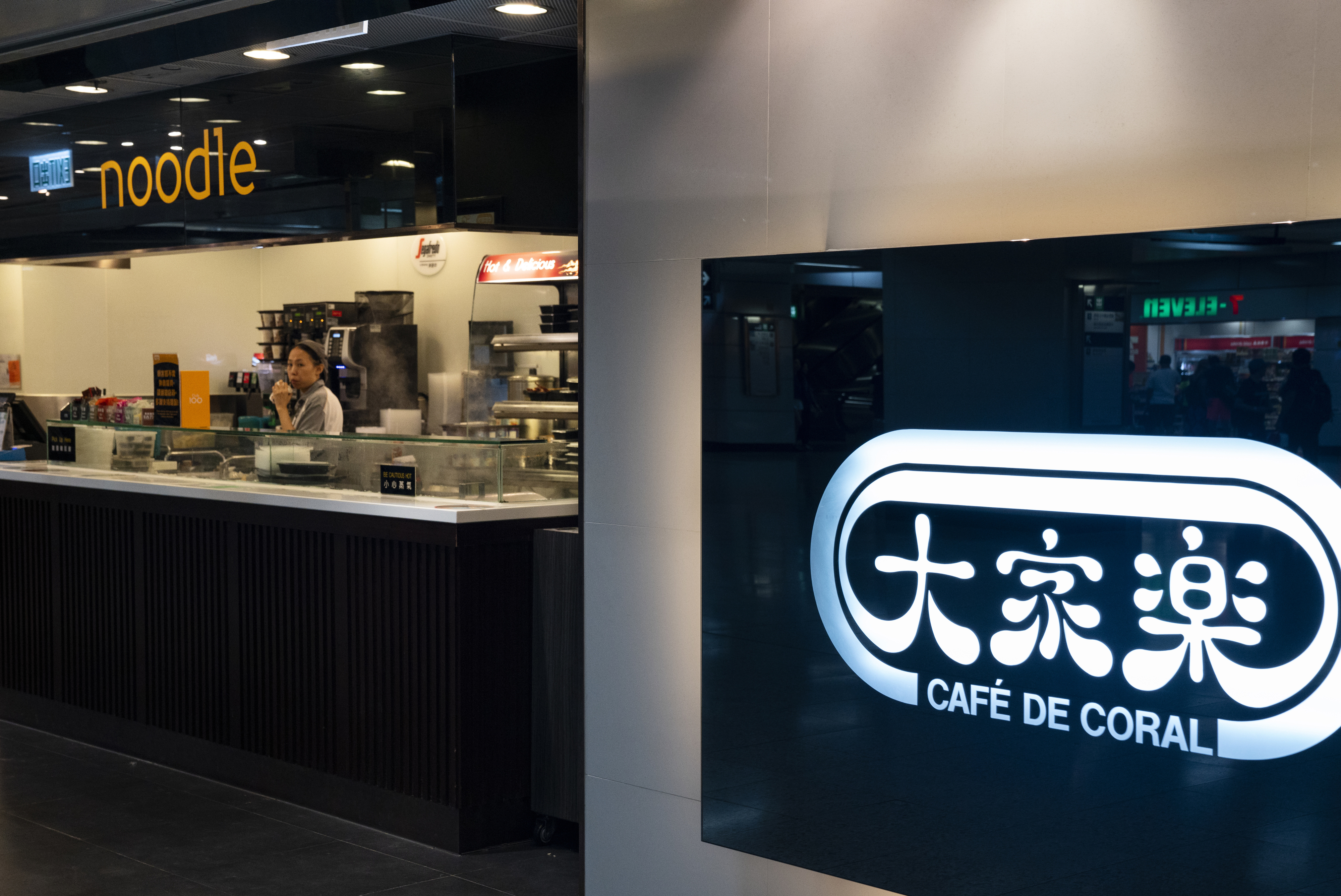 Restaurant signage &quot;Café de Coral&quot; next to a food counter with an employee behind it