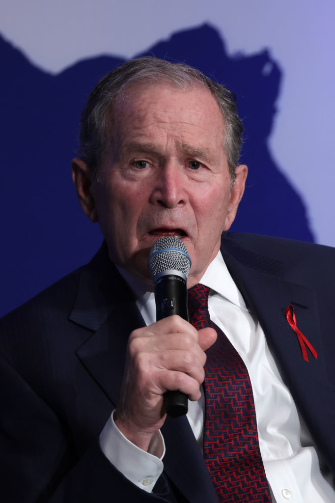 George W. Bush in a suit, speaking into a microphone, with a red ribbon on his lapel