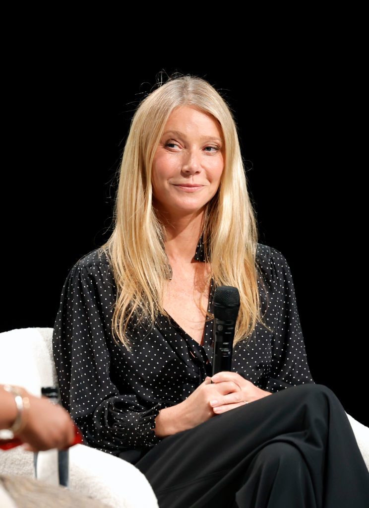 Gwyneth Paltrow seated in an event, wearing a polka dot top and black trousers, holding a microphone