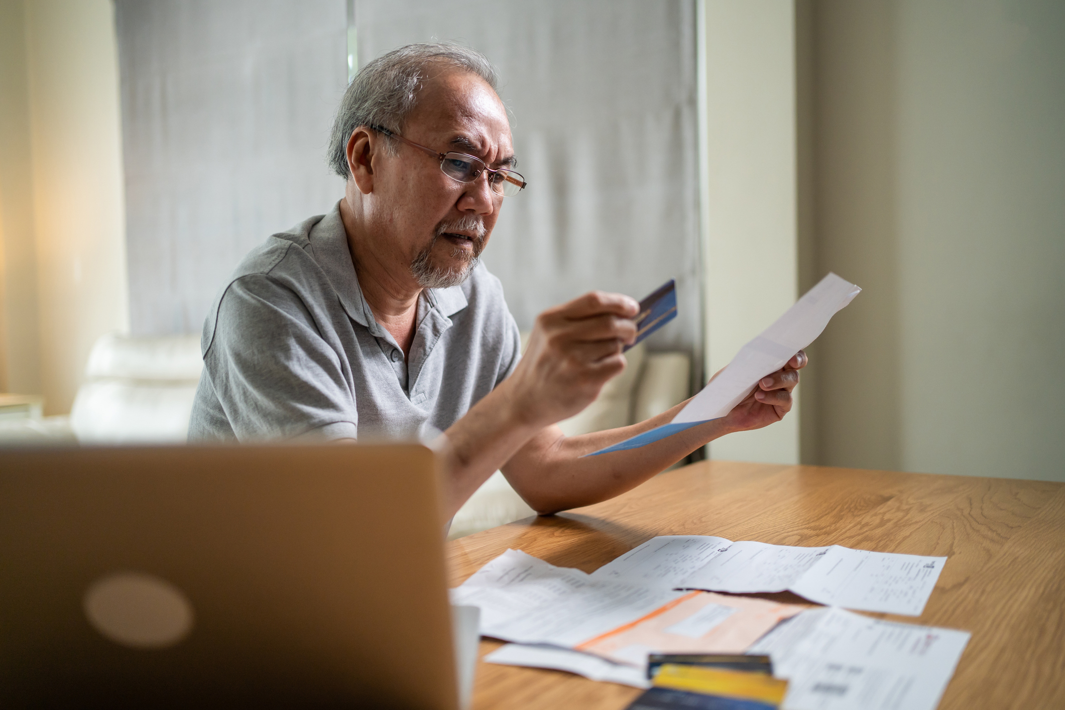 An older adult is holding a credit card and paper, looking confused, with a laptop and documents on the table