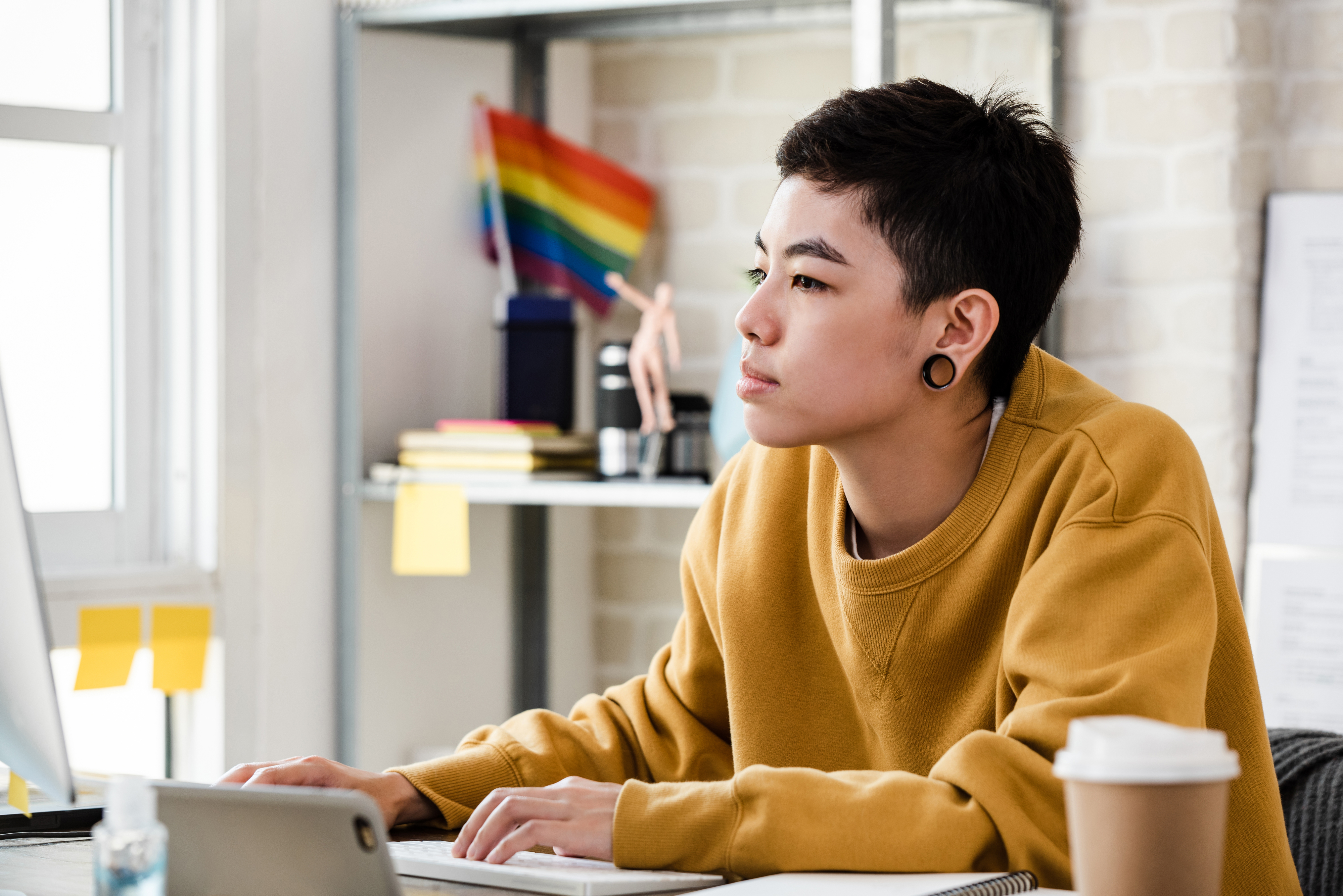 Person in a yellow top working on a computer, with a pride flag in the background