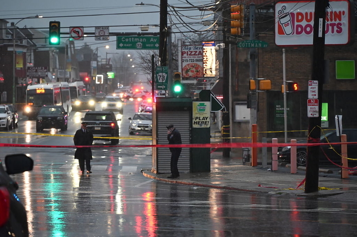 交通規制のテープが張られた通り、警官が現場を見守っている。雨の降る中、店と信号機が写っている。