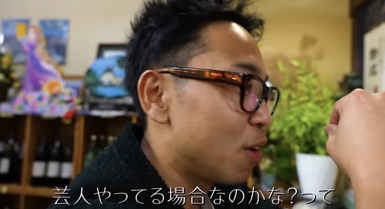 男性が腕を見ながら何かを考えている様子です。背景にはアート作品があります。