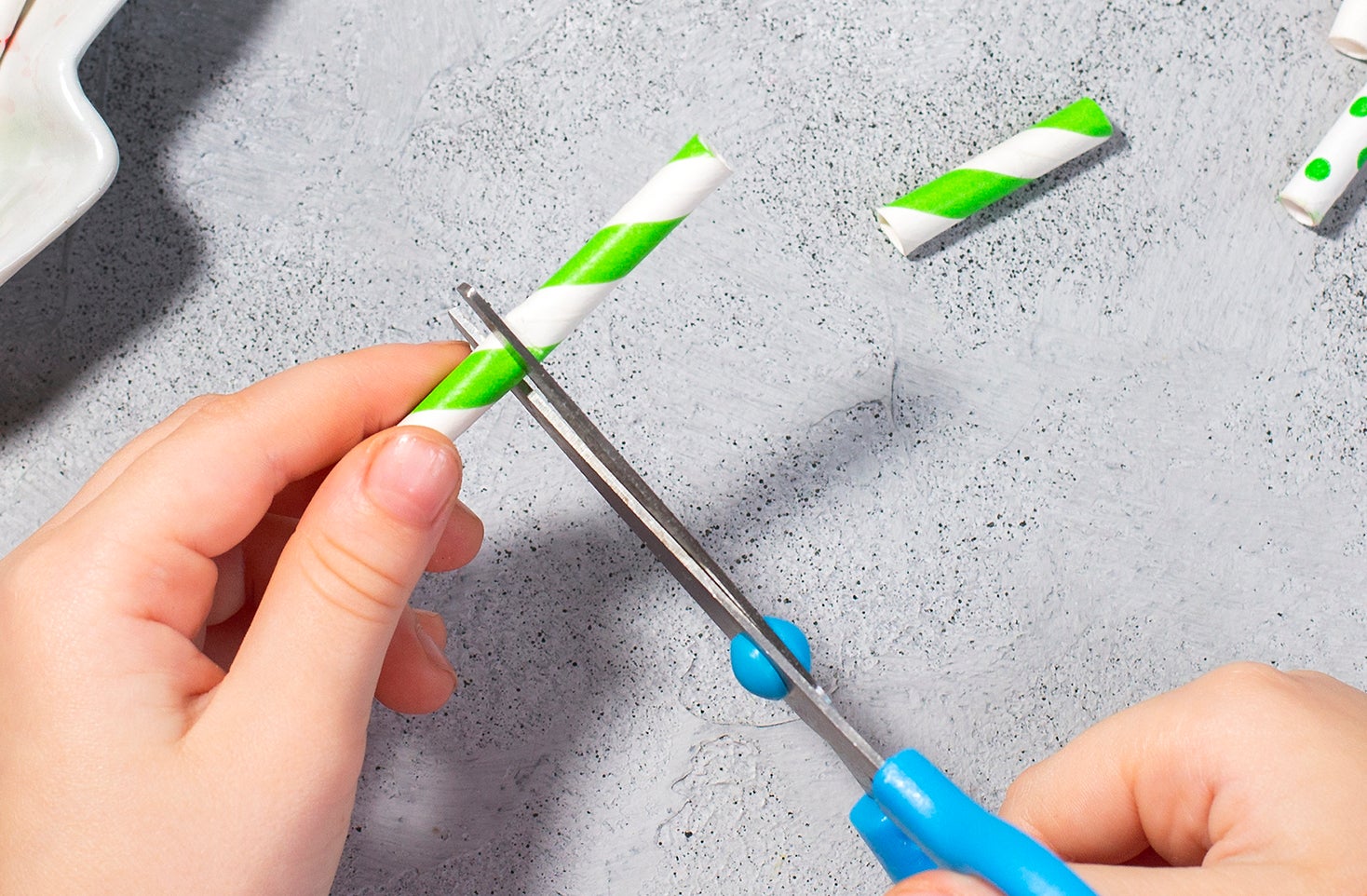 Hands cutting a plastic straw, with various straws and a dish on the table