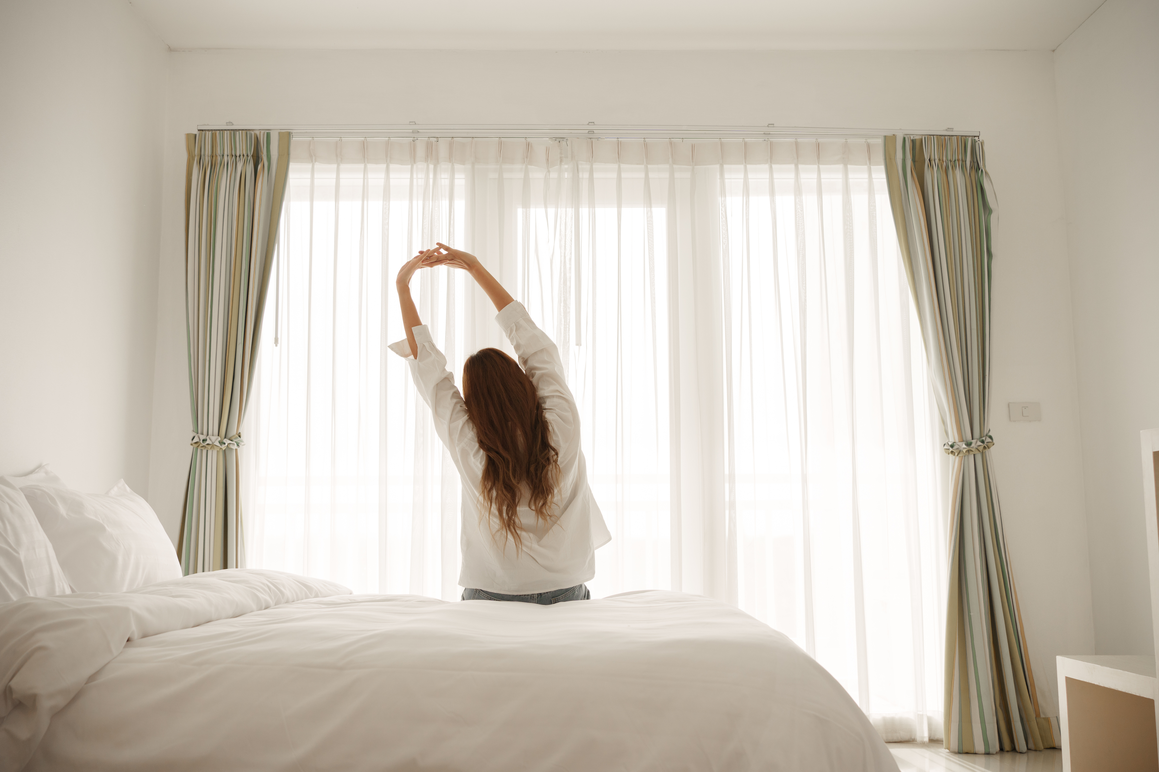 A person stretching while sitting on a bed facing a window with curtains