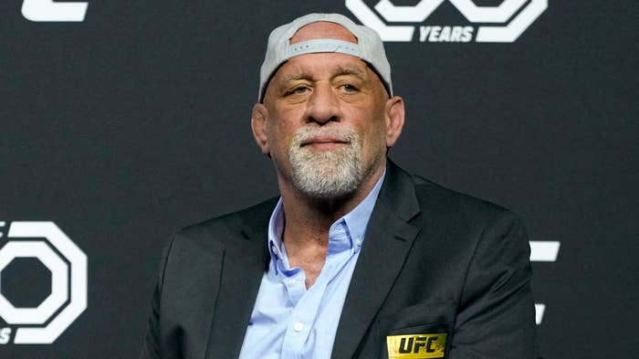 Man in a cap, shirt, and blazer sits in front of a UFC backdrop at a press event