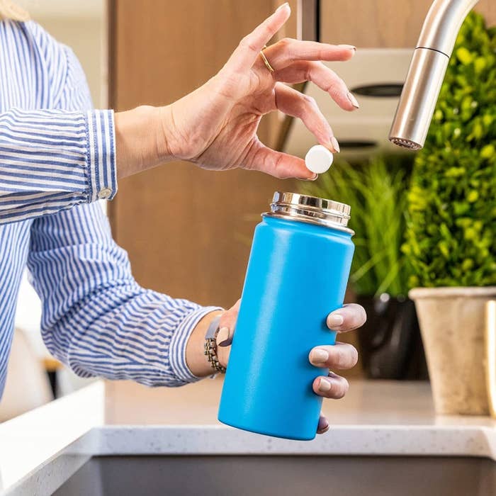 Person dropping a round cleaning tablet into a blue reusable water bottle near a kitchen tap