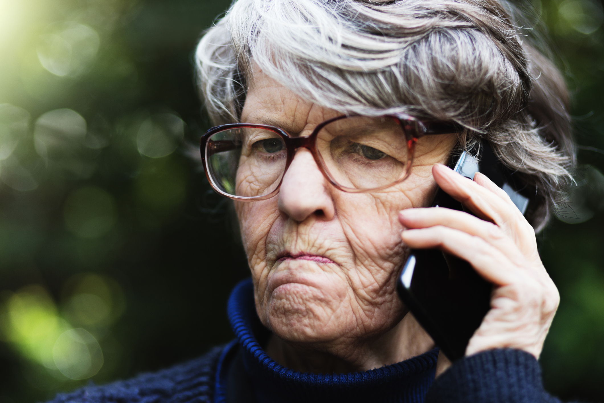 Elderly woman with glasses looks concerned while talking on a cell phone outdoors