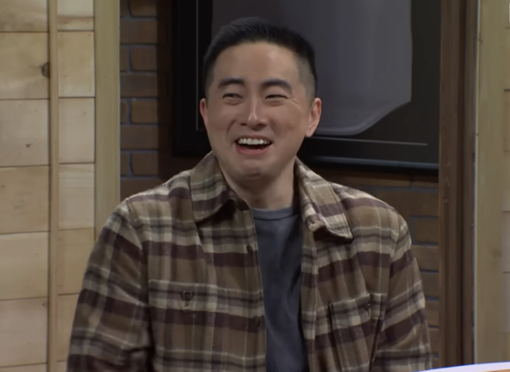 Man in a plaid shirt smiles sitting at a table, in a comedy skit setting
