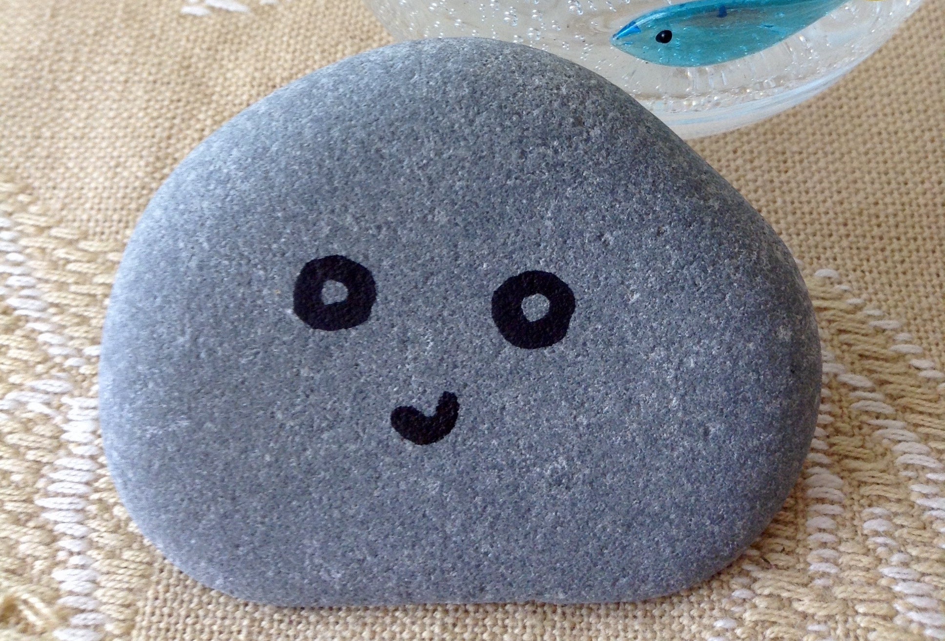 A smooth stone with a drawn-on cute face rests on a textured surface near a clear glass with fish designs