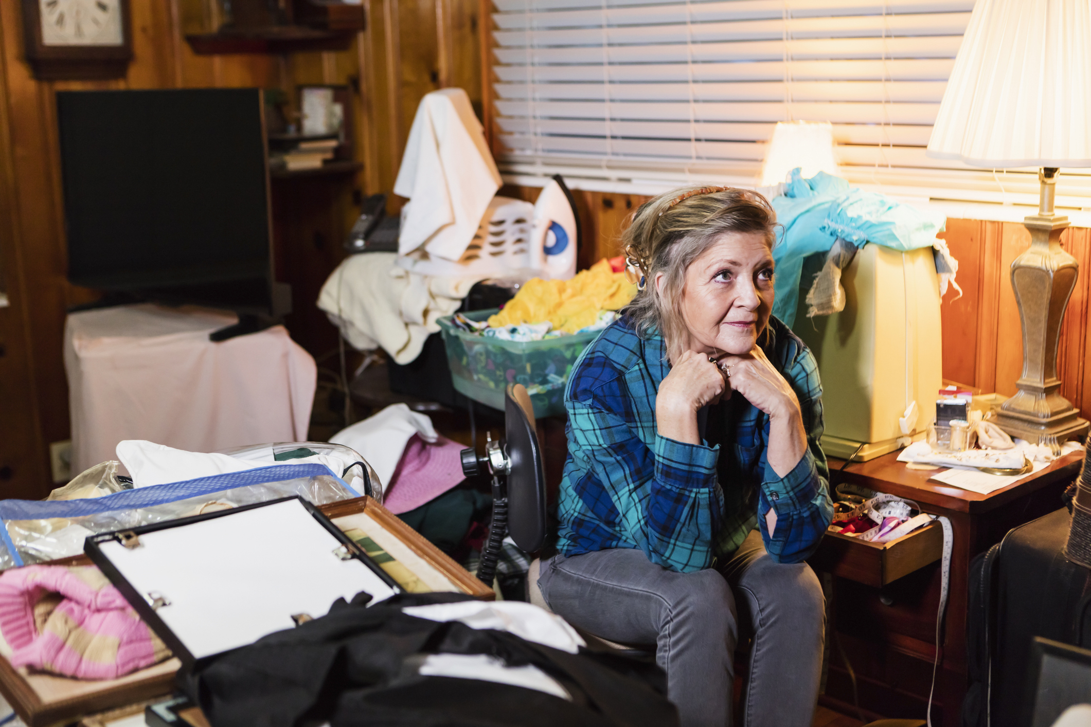 Woman sitting amidst a cluttered room, looking pensive, surrounded by laundry and miscellaneous items