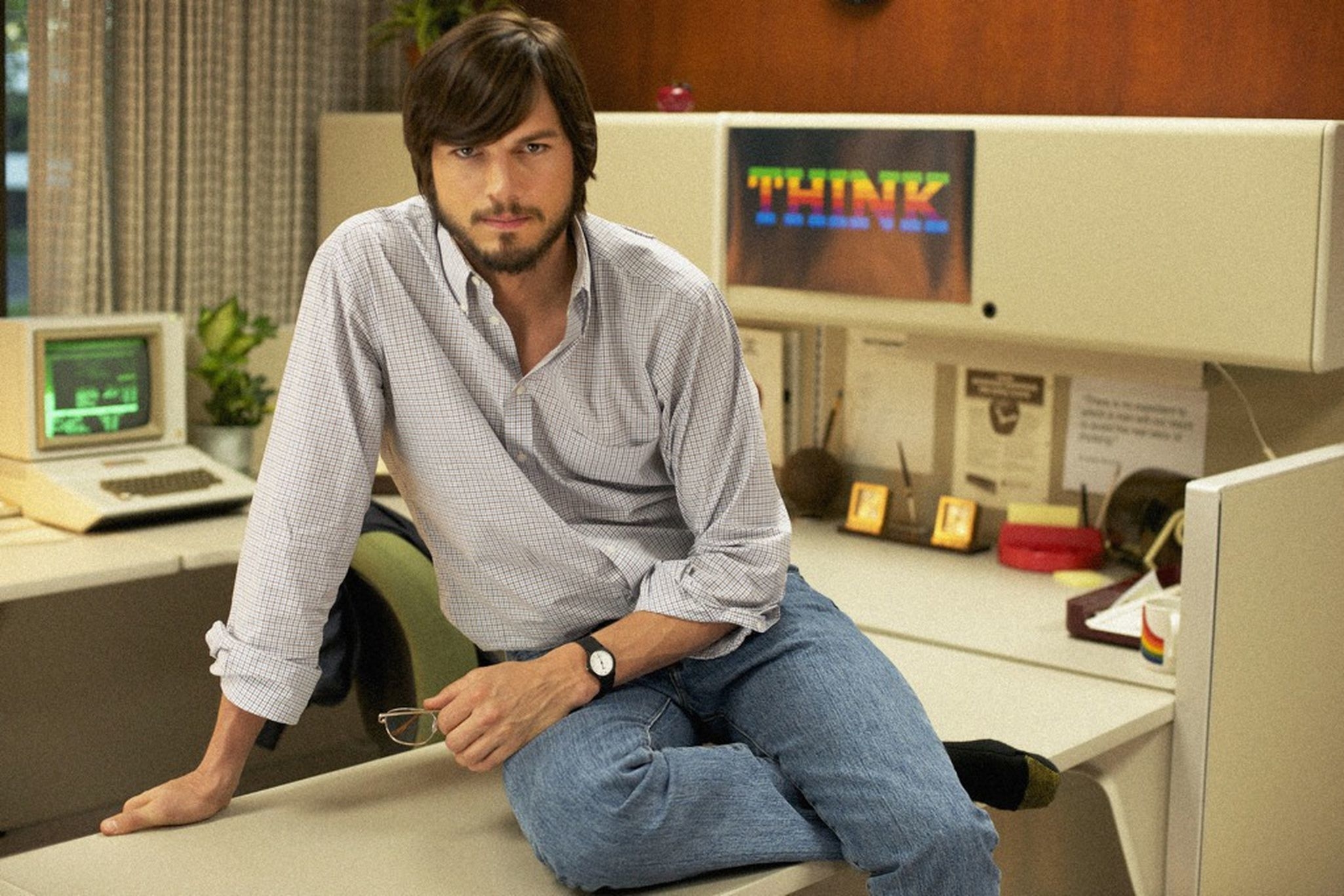 Man resembling Steve Jobs sitting on a desk in an office, with a retro computer and &quot;THINK&quot; poster in background