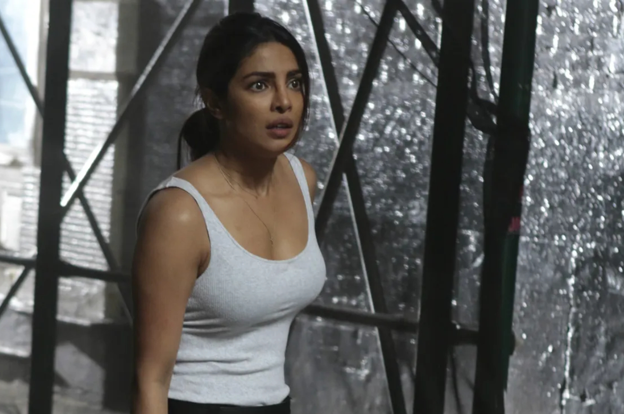 Priyanka Chopra in a white tank top, looking concerned, in a scene with shattered glass in the background