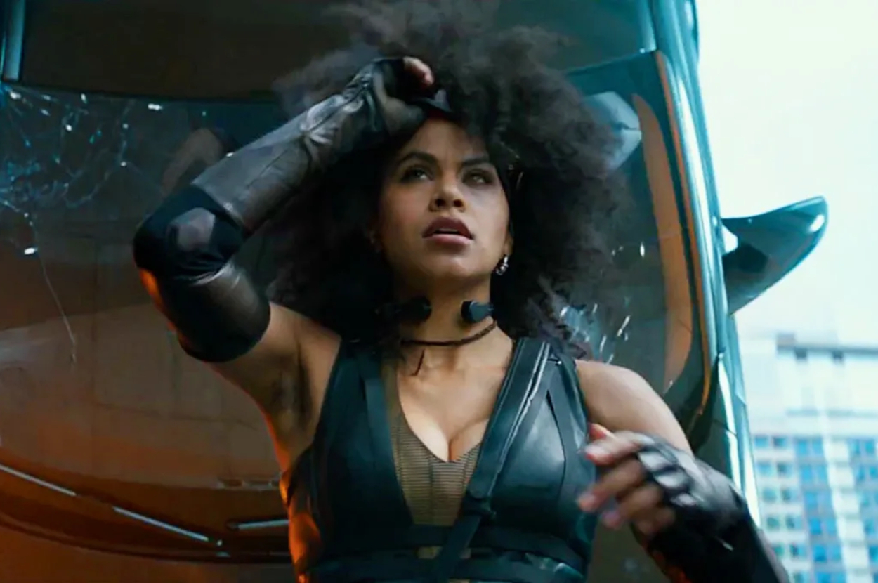 Domino from Deadpool wearing a black outfit, poised for action beside a vehicle