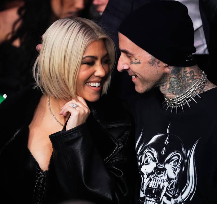 Travis and Kourtney smiling and embracing at an event, she in a black jacket and he in a shirt with skull graphic