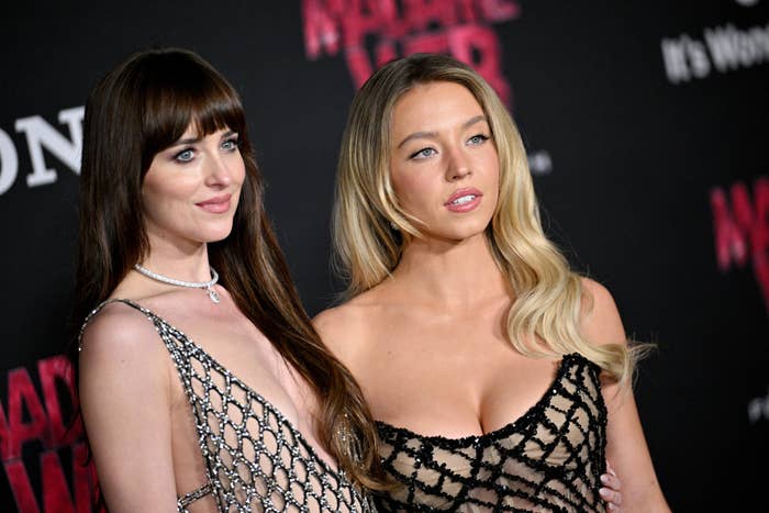 Dakota Johnson and Sydney Sweeney in elegant dresses pose together at an event. Dakota is wearing a chainmail-style dress and Sydney is wearing a lacy, scoop-neck gown