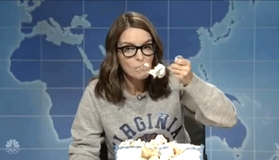 Tina Fey as a comedian on SNL, eating cake from a sheet pan. She wears a gray sweatshirt and glasses