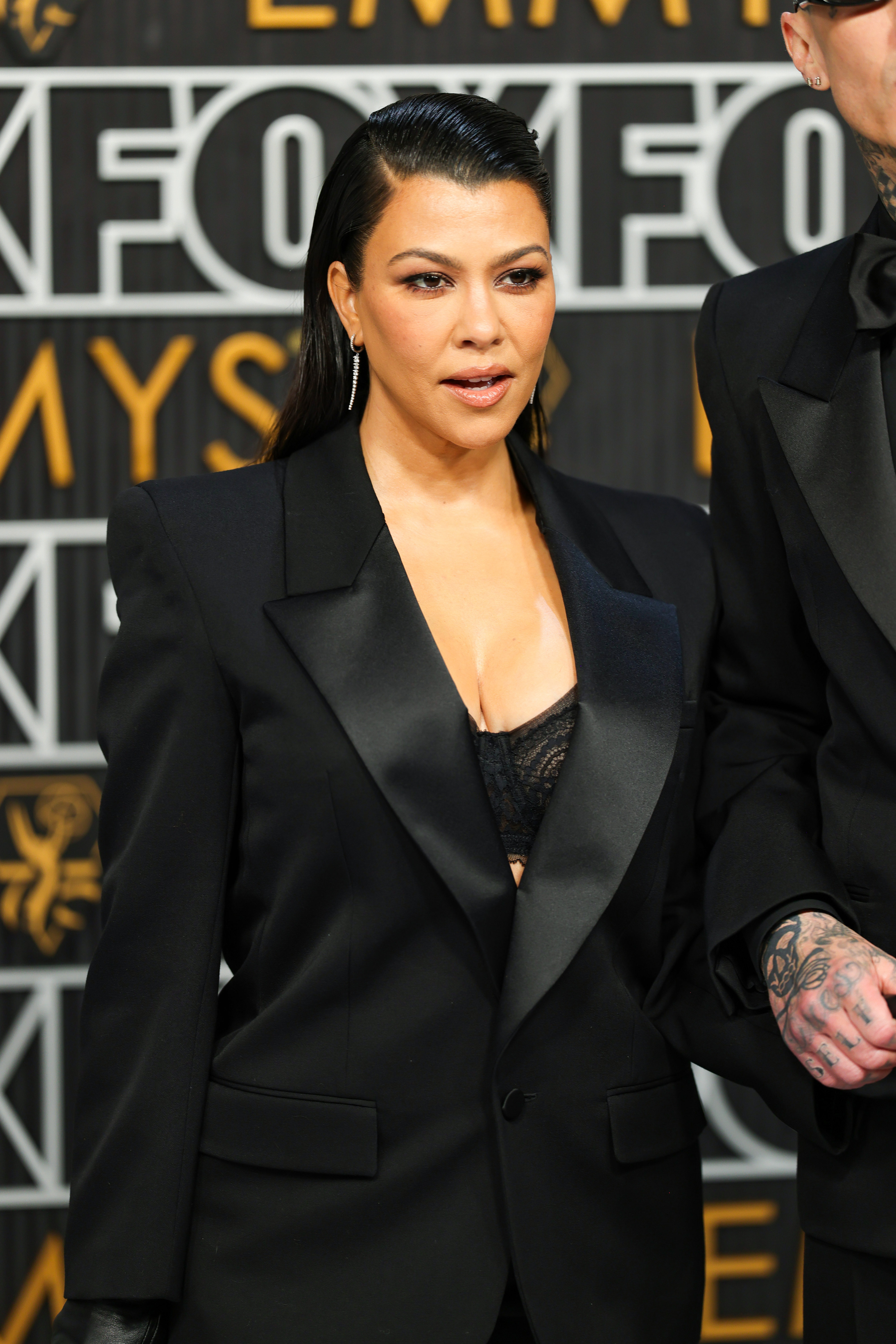 Kourtney in a suit with a plunging neckline, standing next to Travis at a media event