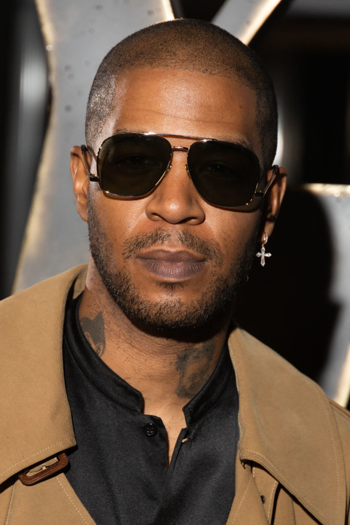 Cudi in sunglasses wearing a trench coat with an earring and tattoos visible on his neck