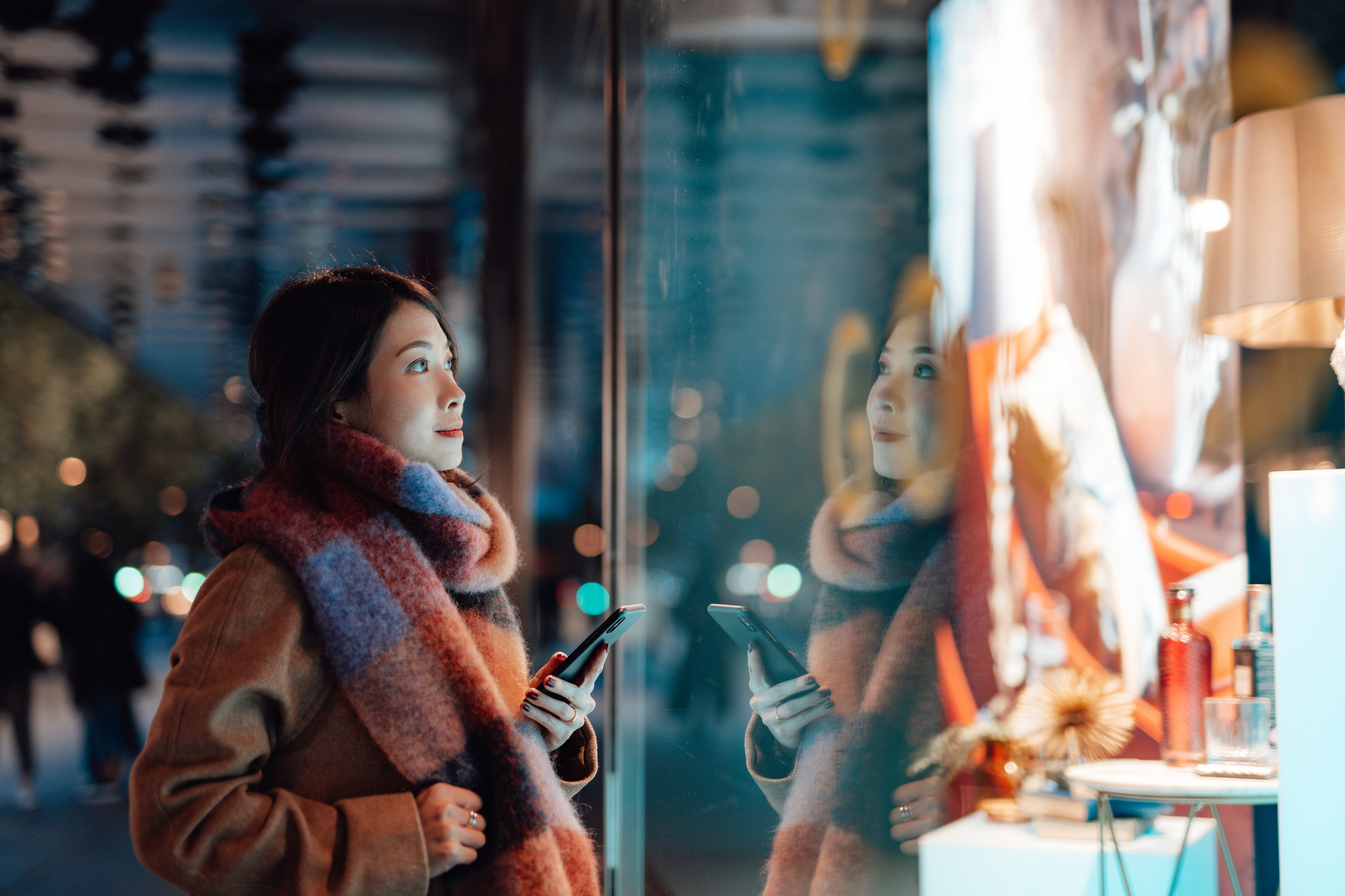 Woman gazing at a window display, wearing a warm scarf, holding a smartphone
