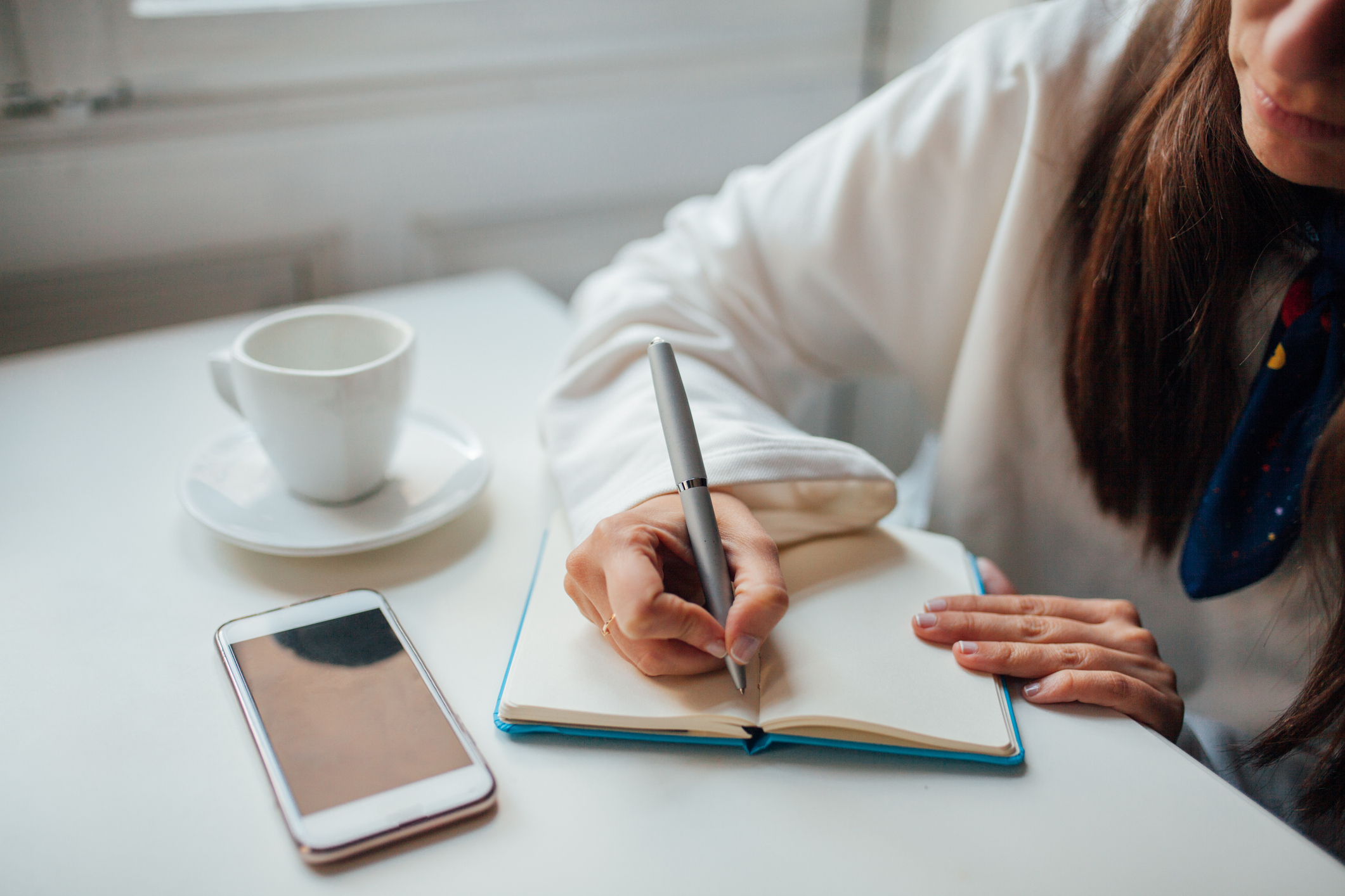 Person writing in a notebook beside a smartphone and coffee cup