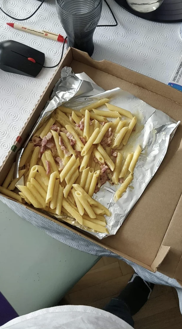 A box of dry-looking penne pasta with pieces of bacon on foil