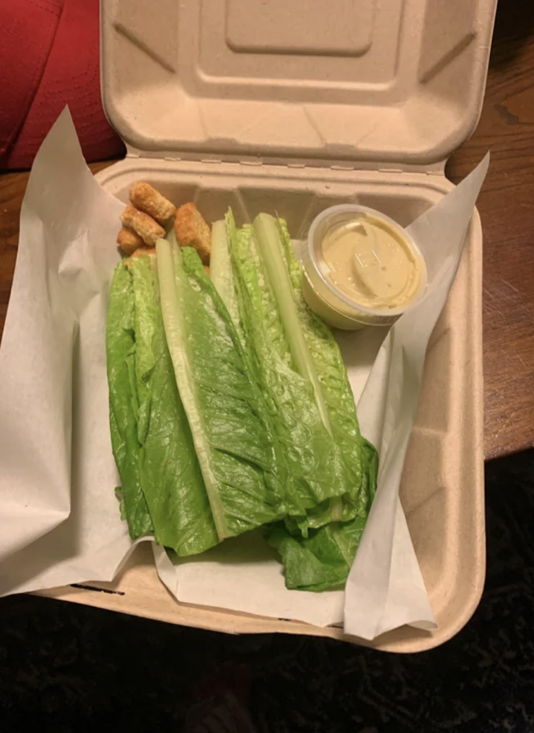 Takeout box with whole lettuce leaves and a side of dipping sauce and a few croutons