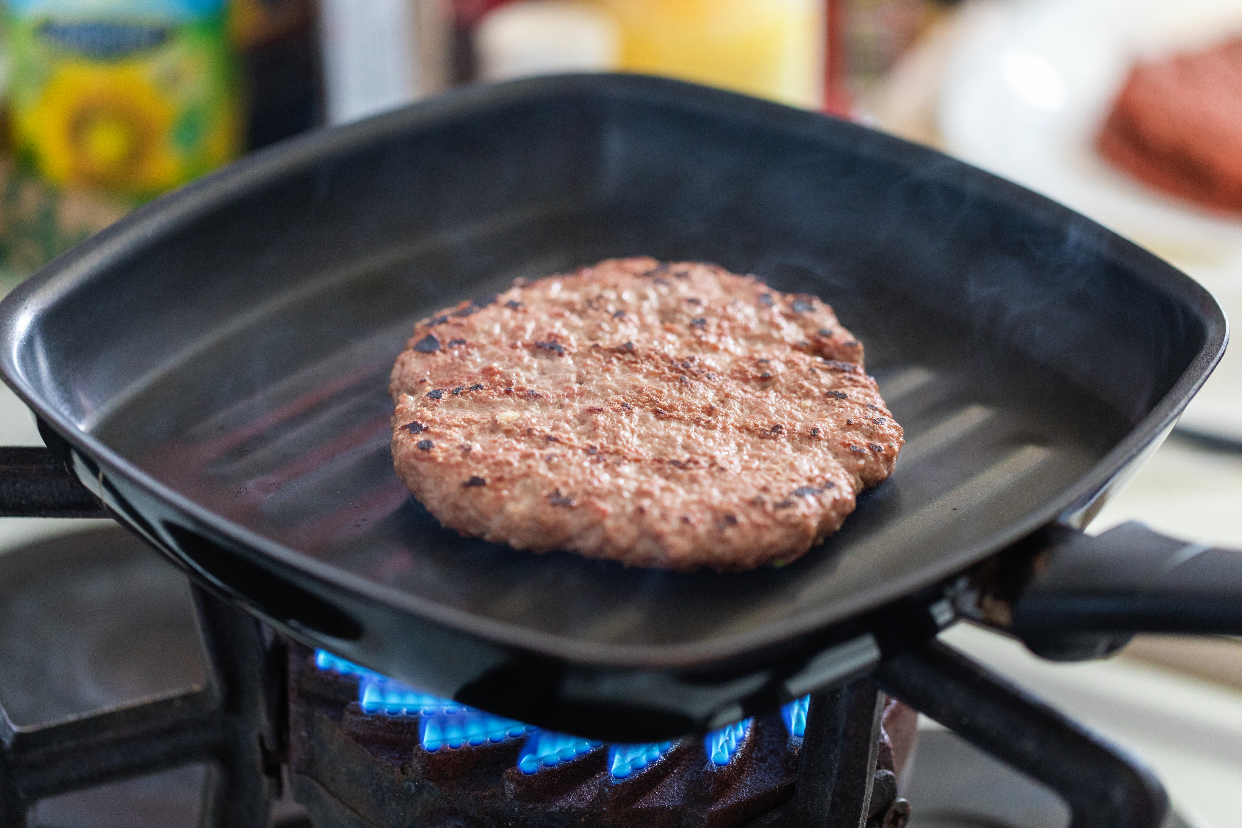 A hamburger patty cooking in a frying pan on a lit gas stove
