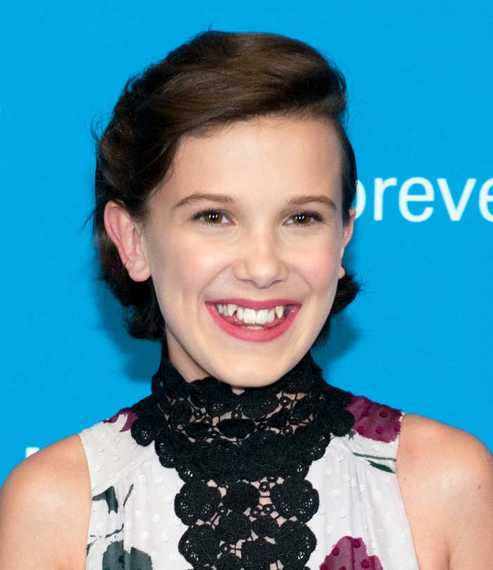 Millie Bobby Brown smiling, wearing a patterned dress with a black lace collar, at an event