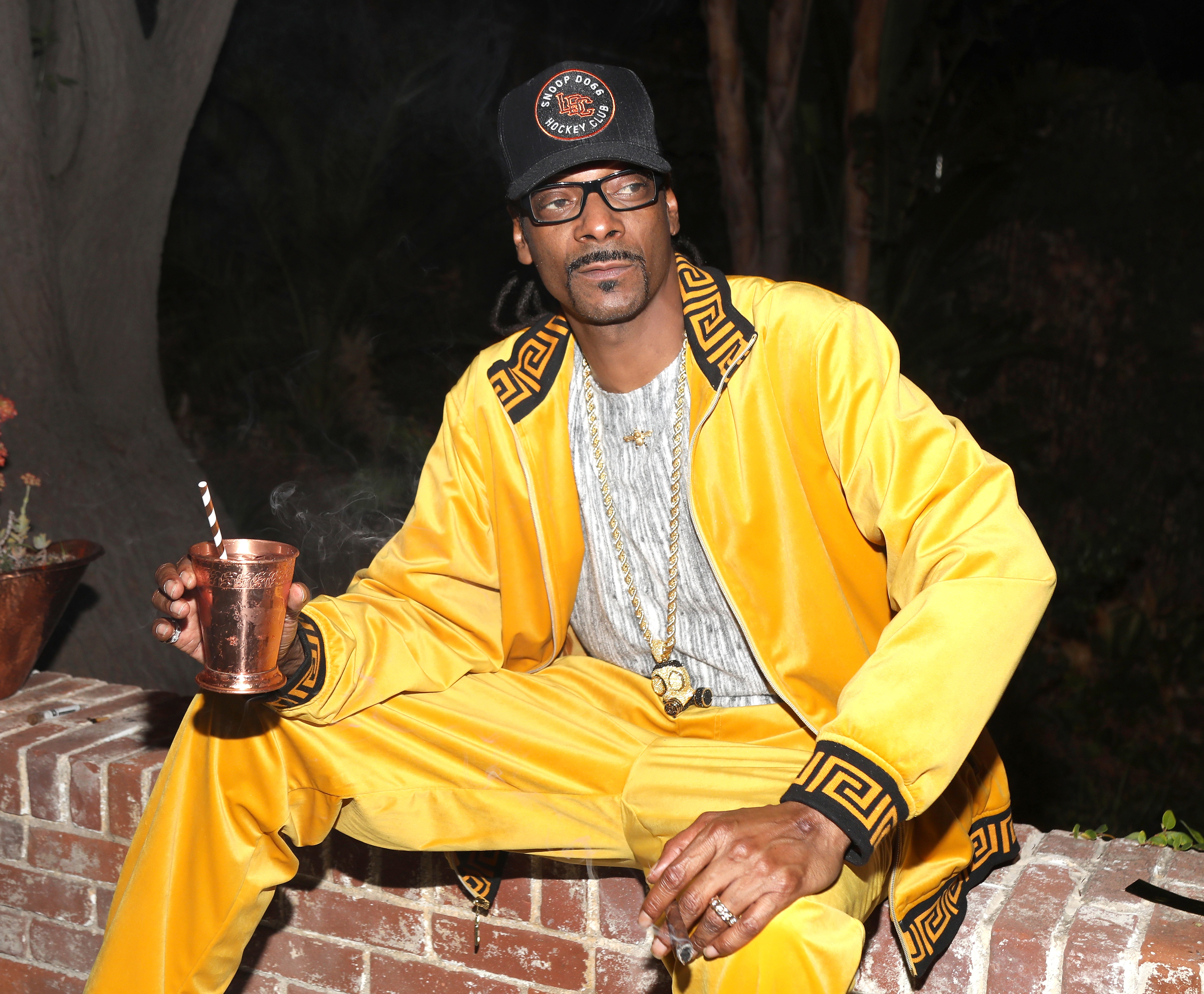 Snoop Dogg sits casually holding a copper mug, wearing a yellow and black tracksuit with intricate designs