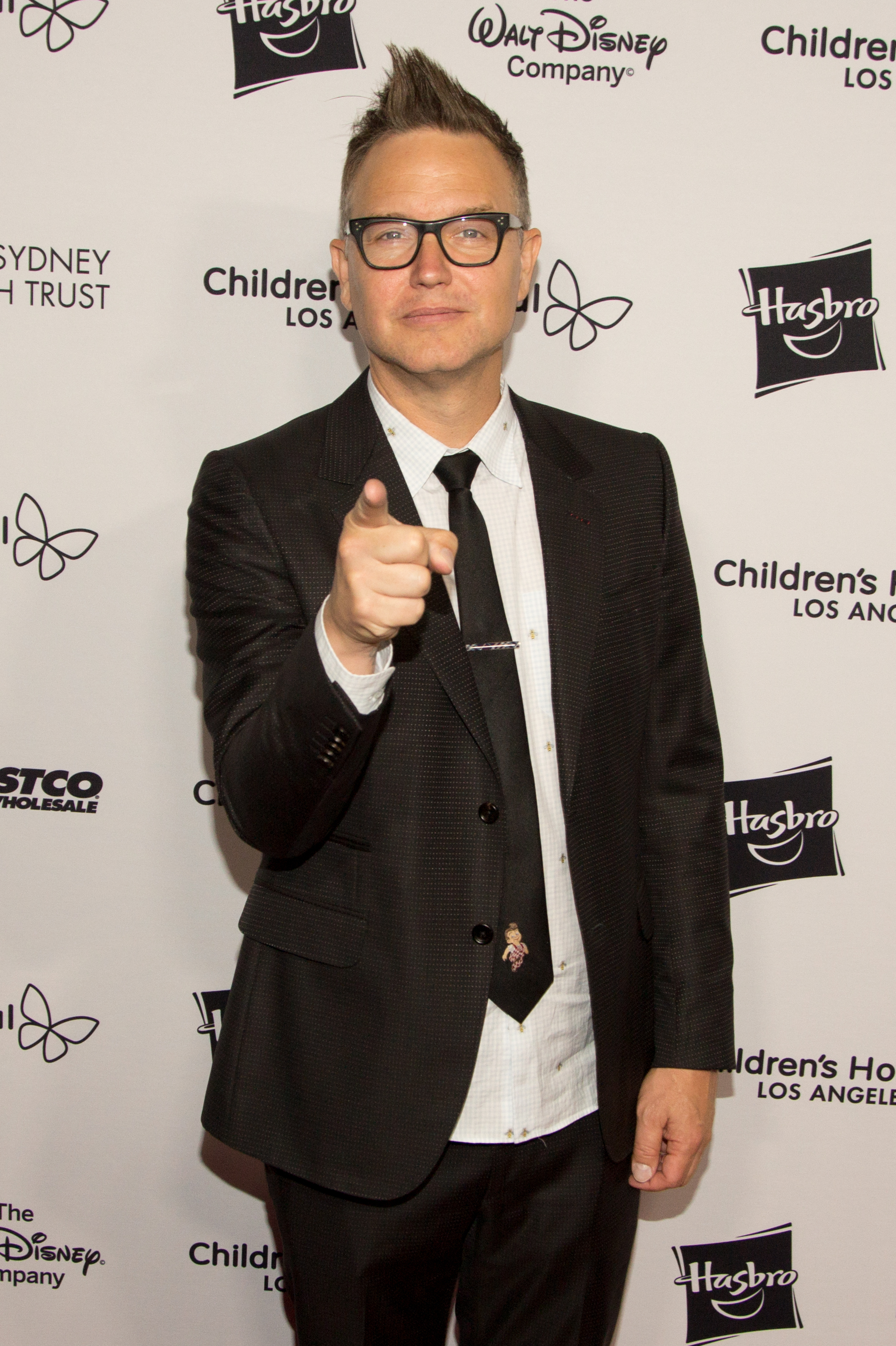 Mark Hoppus in a black suit with a patterned tie giving a thumbs-up at a charity event