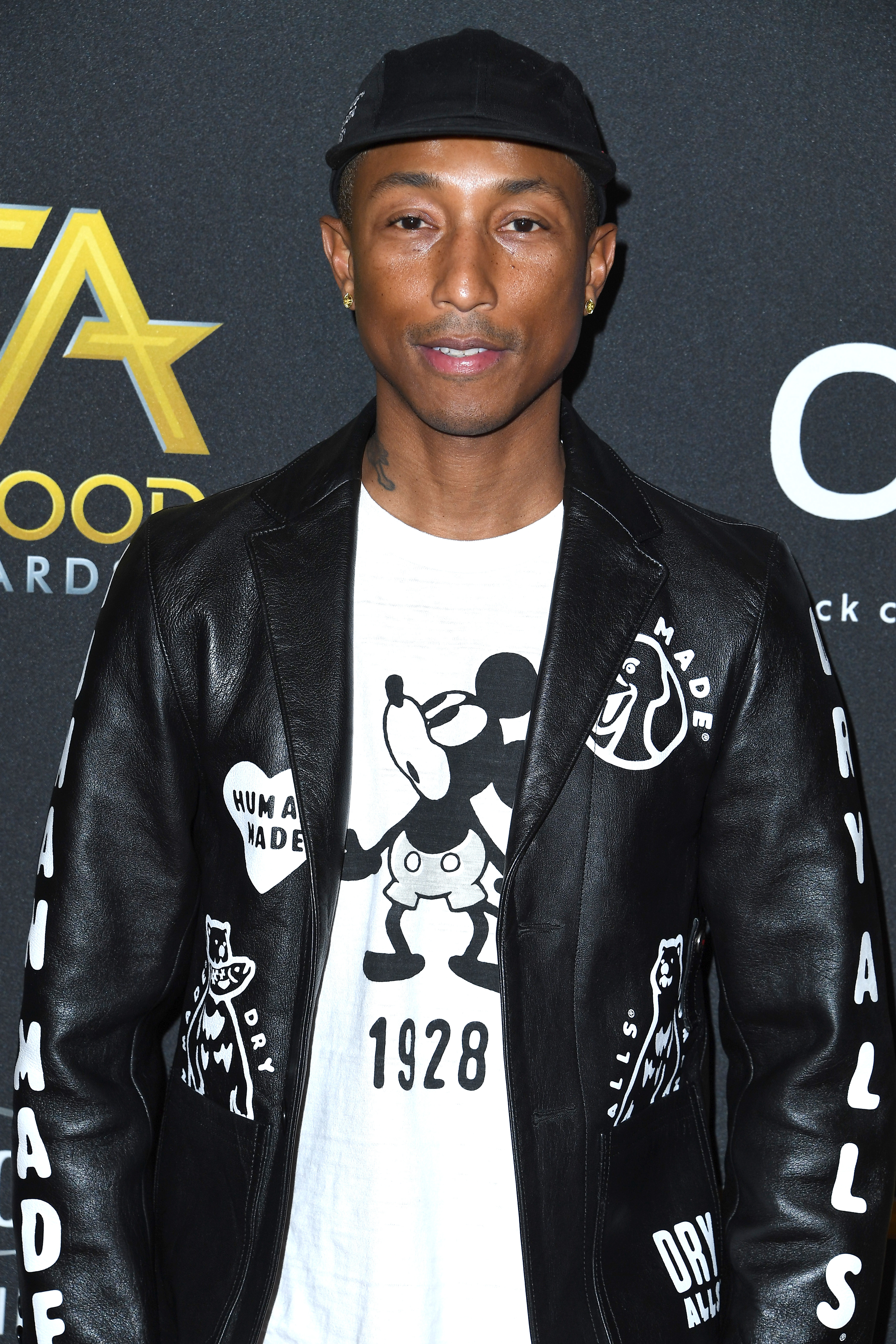 Pharrell Williams in a leather jacket with graphic designs, a tee with Mickey Mouse, and a black hat at an awards event
