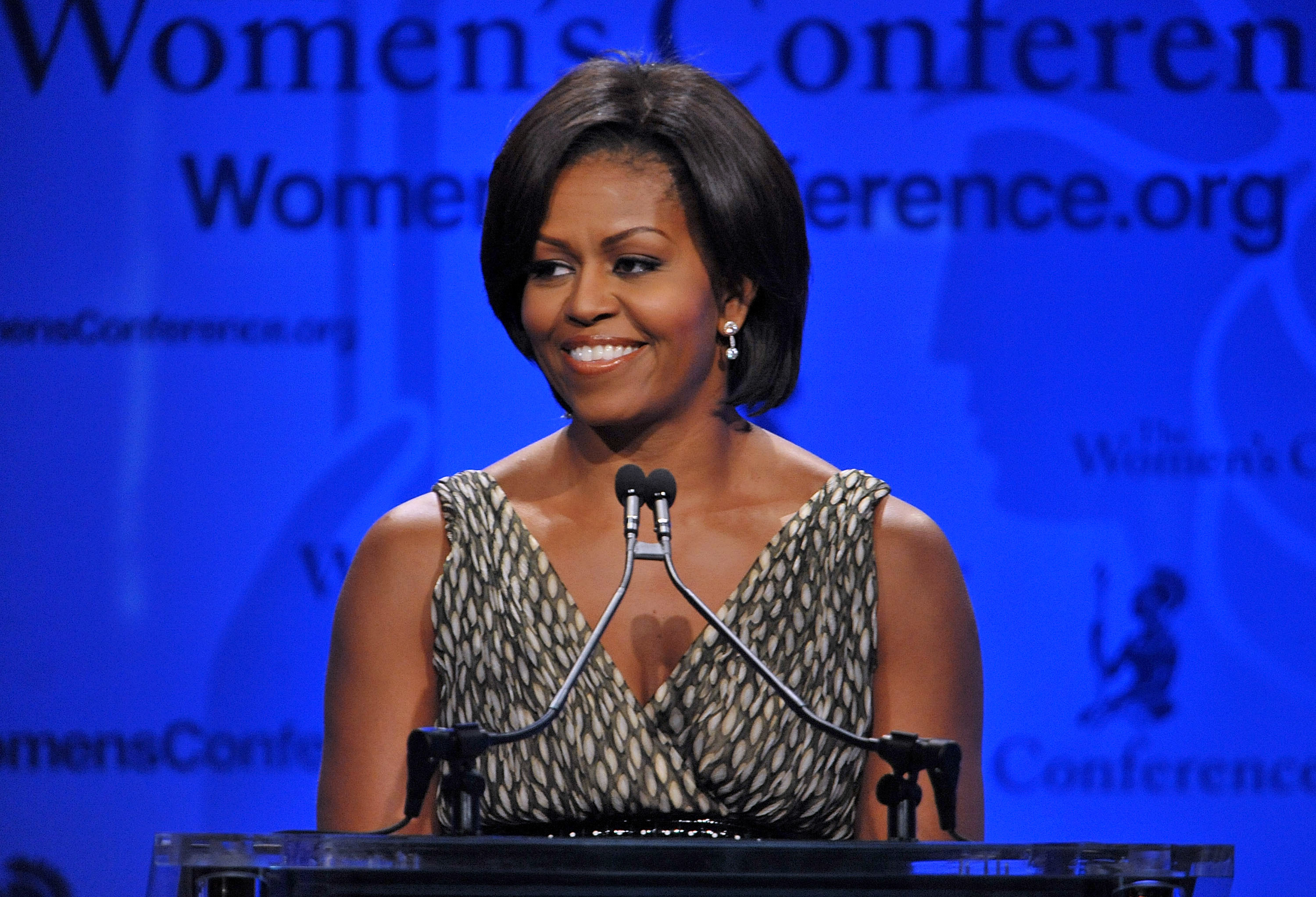 Michelle smiling and standing at a podium with a microphone, addressing an audience, wearing a sleeveless V-neck outfit