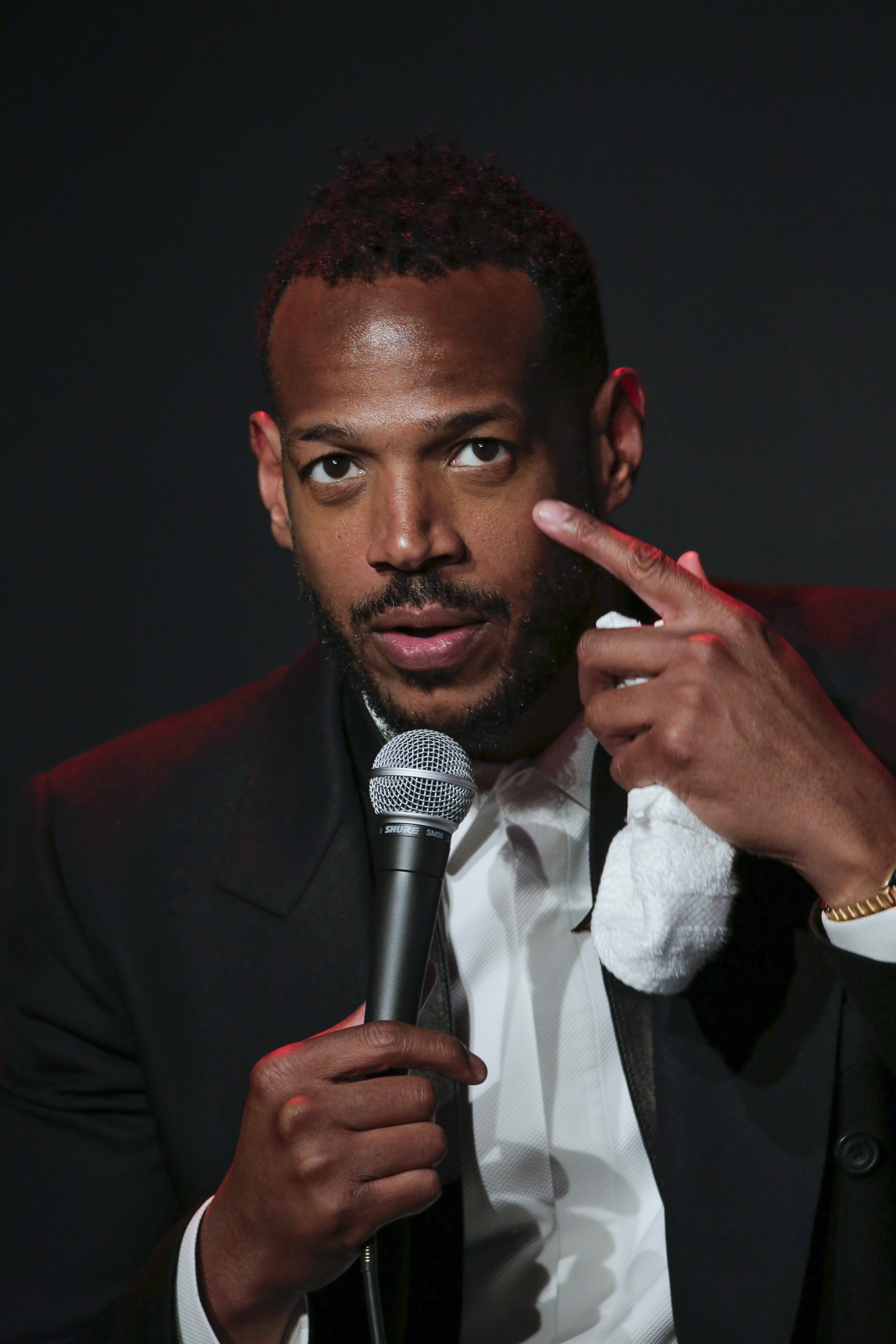 Marlon Wayans in a suit, speaking into a microphone, pointing at his head