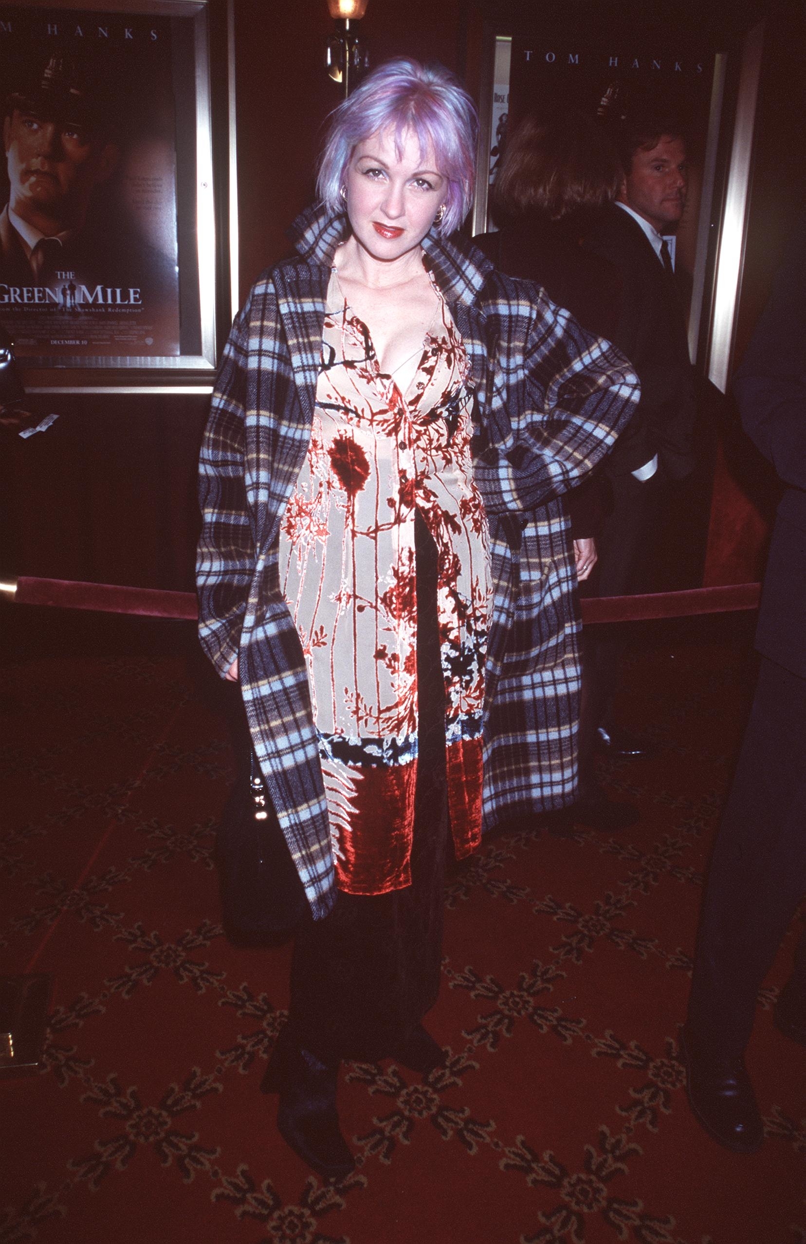 Cyndi on red carpet in plaid coat over patterned dress, standing near a movie poster