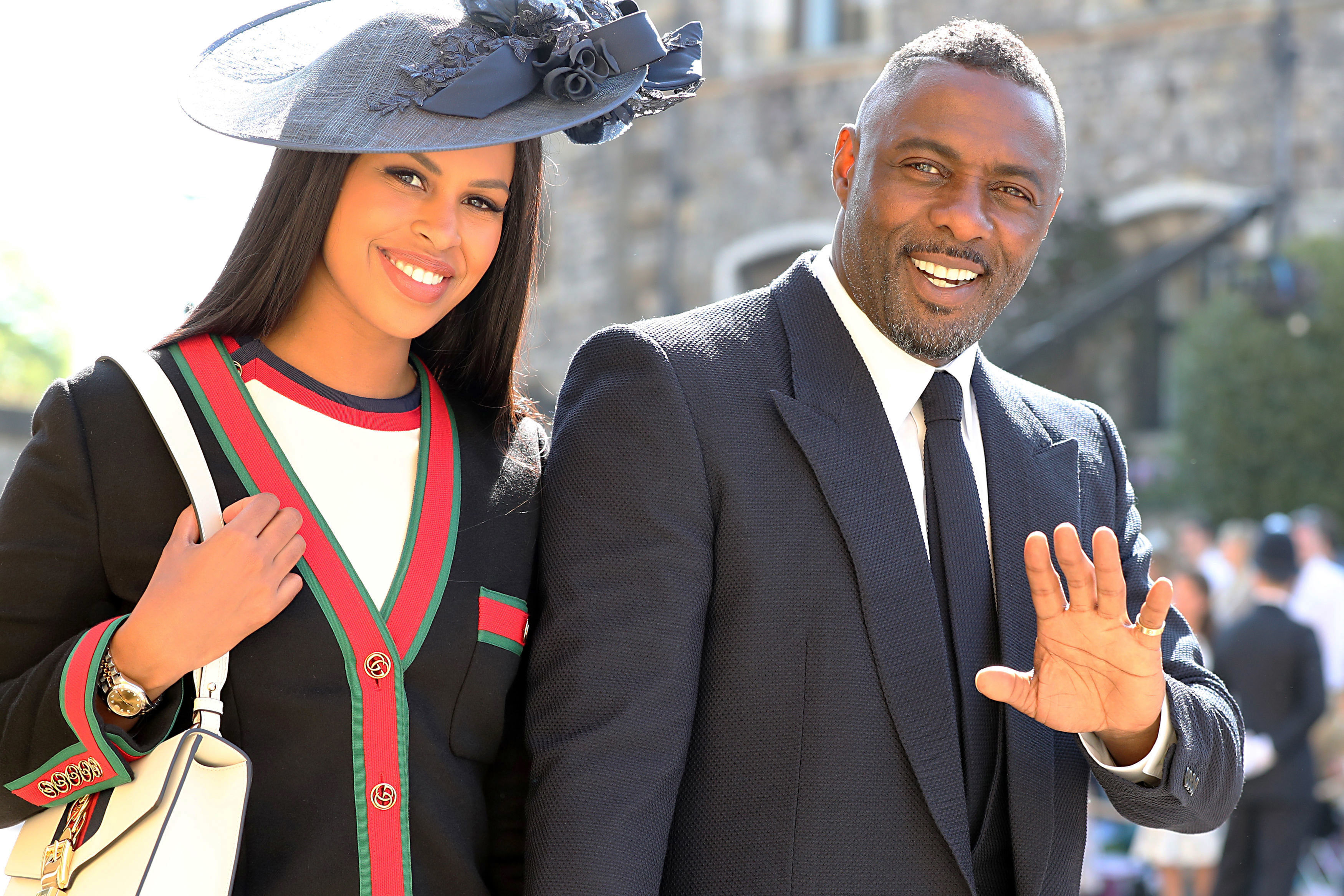 Sabrina Dhowre Elba in hat and striped outfit and Idris in suit, both smiling and waving
