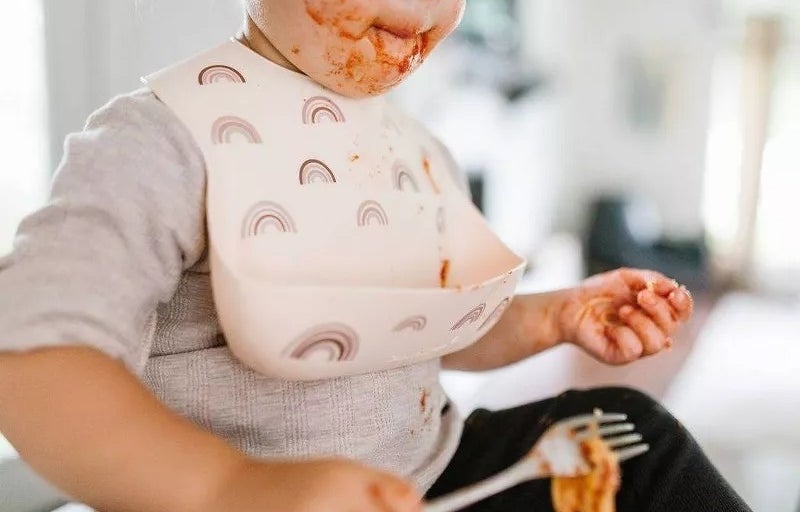 Toddler with food on face holding a fork, wearing a bib with rainbow pattern in a high chair