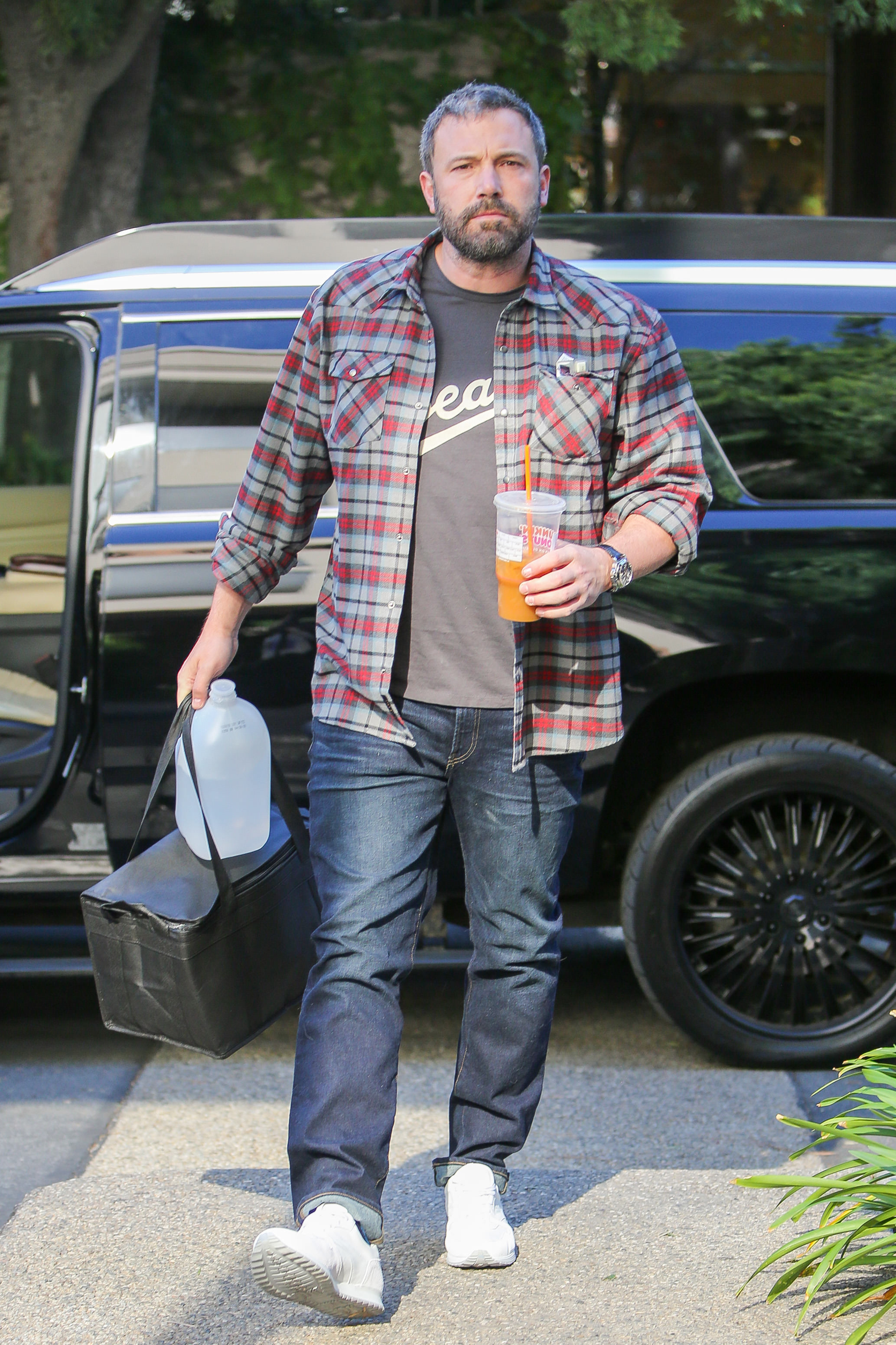 Ben in a plaid shirt over a graphic tee, flannel shirt, denim, and sneakers, carrying a Dunkin&#x27; coffee and a bag, exiting a vehicle