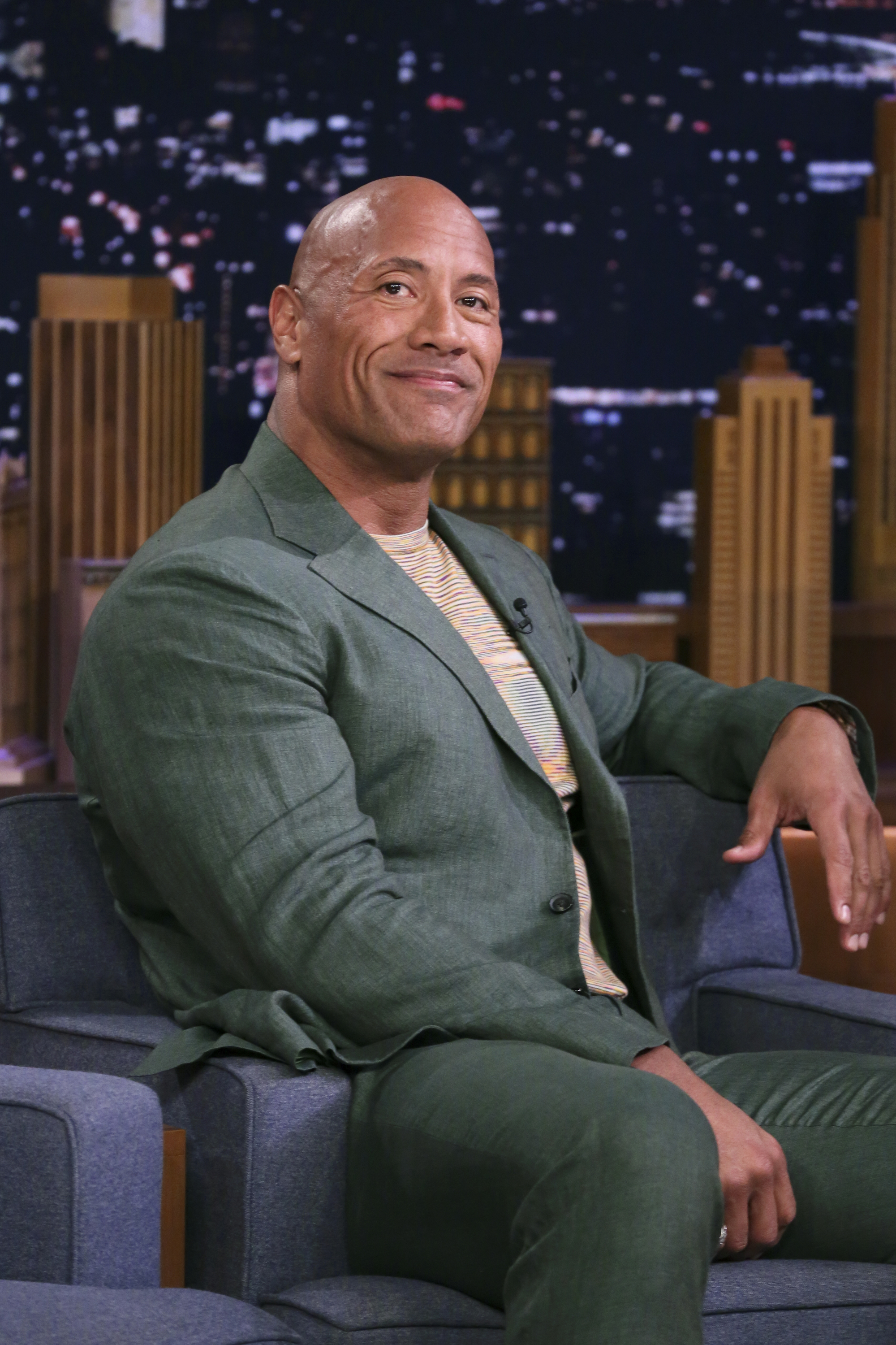 Dwayne Johnson smiles, seated in a talk show setting, wearing a green suit with a textured shirt