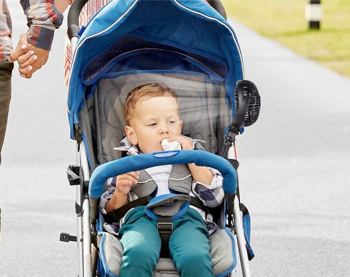Toddler model in a stroller with an attached fan, adults alongside, illustrating a portable stroller fan product