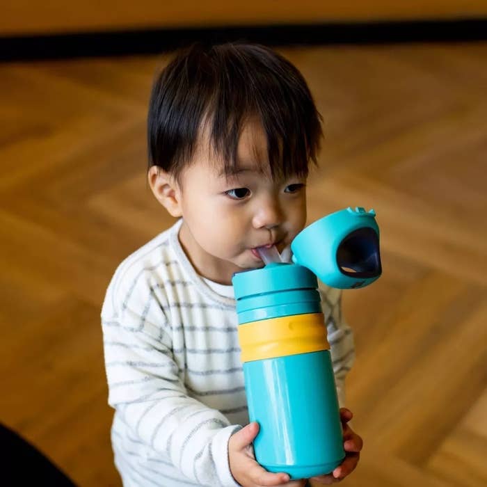 Child holding a turquoise and yellow sippy cup, sipping from the straw, wearing a striped long-sleeve shirt